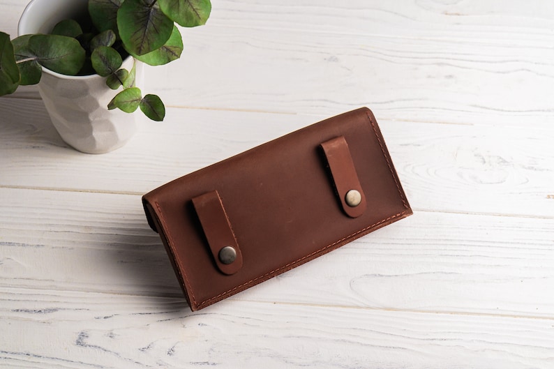 May include: A brown leather wallet with two snap closures. The wallet is lying on a white wooden surface.