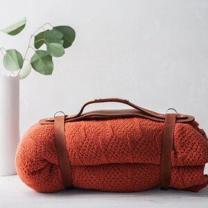 May include: A rolled-up orange blanket with a brown leather handle and straps. A white vase with green leaves is to the left of the blanket. The blanket is made of a knitted material.