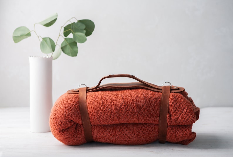 May include: A rolled-up orange blanket with a brown leather handle and straps. A white vase with green leaves is to the left of the blanket. The blanket is made of a knitted material.