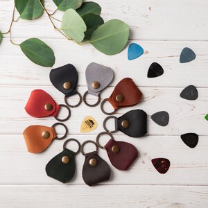 May include: A set of ten leather guitar picks in various colors, including red, brown, green, black, and gray. Each pick has a small loop for attaching to a keychain. The picks are arranged in a circle on a white wooden surface.