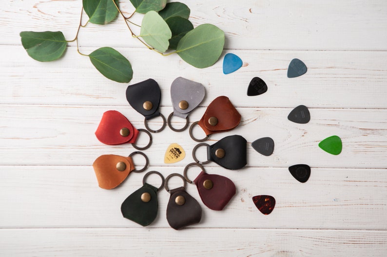 May include: A set of ten leather guitar picks in various colors, including red, brown, green, black, and gray. Each pick has a small loop for attaching to a keychain. The picks are arranged in a circle on a white wooden surface.