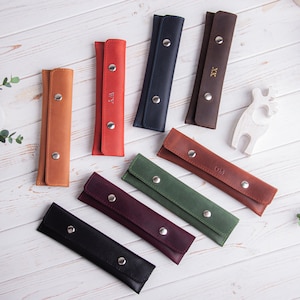 May include: Nine leather pencil cases in various colors, including brown, red, green, blue, and black. Each case has a silver snap closure and is personalized with initials.