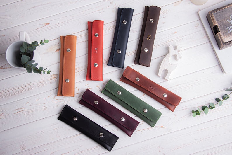 May include: Nine leather pencil cases in various colors, including brown, red, green, blue, and black. Each case has a silver snap closure and is personalized with initials.
