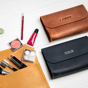 May include: An assortment of makeup products and leather cosmetic bags. The bags are brown and navy blue, with the names "CAMBER" and "DOLIE" embossed on them. Makeup items include blush, lipstick, lip gloss, and eyeshadow.
