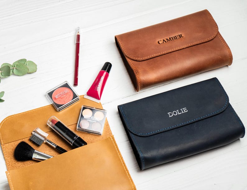 May include: An assortment of makeup products and leather cosmetic bags. The bags are brown and navy blue, with the names "CAMBER" and "DOLIE" embossed on them. Makeup items include blush, lipstick, lip gloss, and eyeshadow.