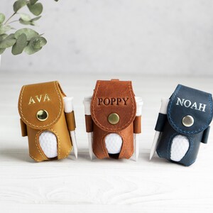 May include: Three personalized golf ball holders in tan, brown, and navy blue leather. Each holder has a golf ball and tee, with names "AVA", "POPPY", and "NOAH" embossed on the front. A decorative plant in a white vase is in the background.