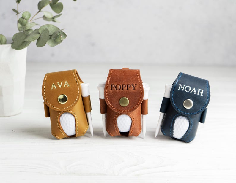 May include: Three personalized golf ball holders in tan, brown, and navy blue leather. Each holder has a golf ball and tee, with names "AVA", "POPPY", and "NOAH" embossed on the front. A decorative plant in a white vase is in the background.