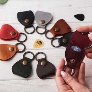 May include: Assortment of eight leather guitar pick holders with key rings. Each holder is a different color, including brown, red, green, black, gray, and burgundy. One holder is open to show a guitar pick inside.