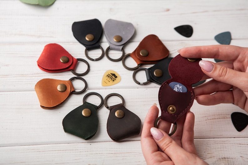 May include: Assortment of eight leather guitar pick holders with key rings. Each holder is a different color, including brown, red, green, black, gray, and burgundy. One holder is open to show a guitar pick inside.