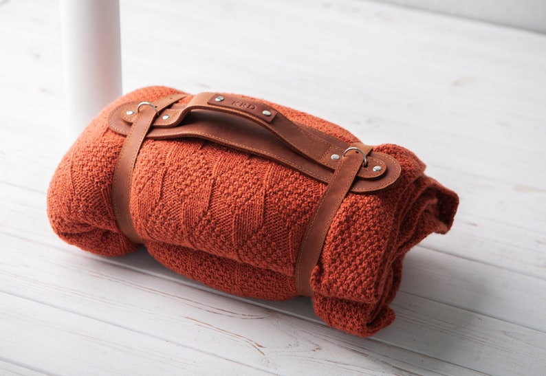 May include: A rolled-up orange blanket with a brown leather carrying strap. The strap has a handle and is secured with metal rivets. The blanket is made of a textured knit fabric. The initials "KPD" are visible on the handle.