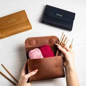 May include: A brown leather knitting needle case with two balls of yarn, one pink and one red, and a set of knitting needles inside. The case is open and the needles are being held by a hand. The case has the name "Alison" embossed on the front.