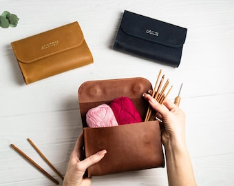 May include: A brown leather knitting needle case with two balls of yarn, one pink and one red, and a set of knitting needles inside. The case is open and the needles are being held by a hand. The case has the name "Alison" embossed on the front.