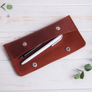 May include: A brown leather pencil case with two pens inside. The case is open and the pens are visible. The case is on a white wooden surface.
