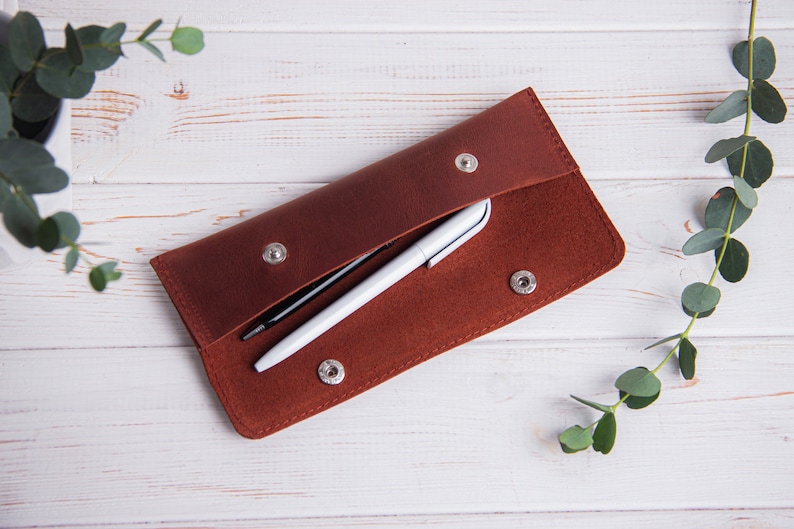May include: A brown leather pencil case with two pens inside. The case is open and the pens are visible. The case is on a white wooden surface.