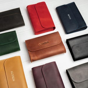May include: A collection of leather wallets in various colors, including black, red, brown, and green. Each wallet features a flap closure and some have personalized text. The wallets are arranged on a white surface.