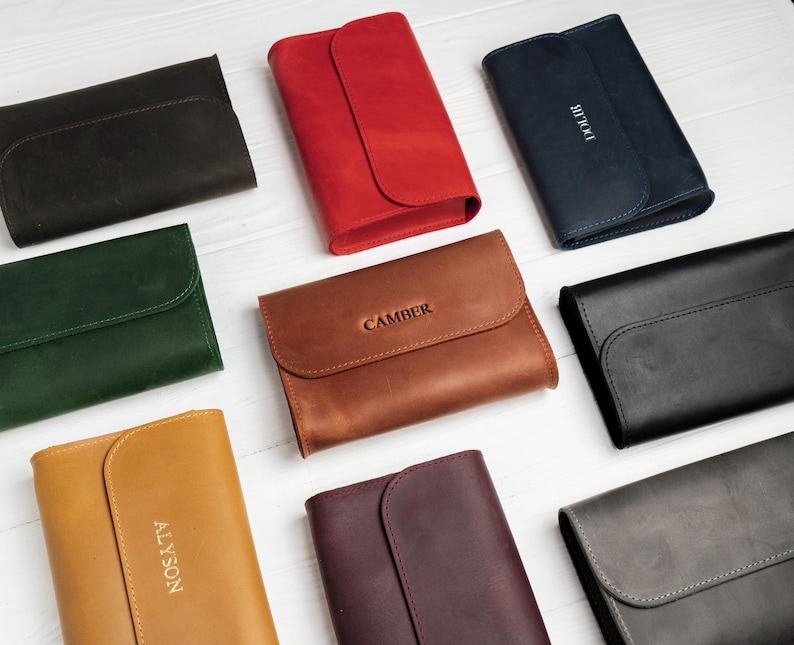 May include: A collection of leather wallets in various colors, including black, red, brown, and green. Each wallet features a flap closure and some have personalized text. The wallets are arranged on a white surface.