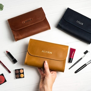 May include: Three leather cosmetic bags in brown, navy, and tan, each with a name embossed on the flap. Makeup items like lipstick, brushes, and eyeshadow are arranged around the bags on a white surface.