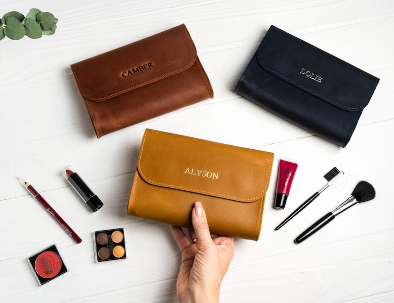 May include: Three leather cosmetic bags in brown, navy, and tan, each with a name embossed on the flap. Makeup items like lipstick, brushes, and eyeshadow are arranged around the bags on a white surface.