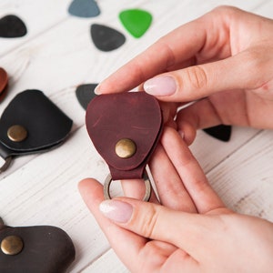 May include: A set of four leather guitar pick holders with keychains. The holders are in brown, black, and burgundy. The keychains are silver.