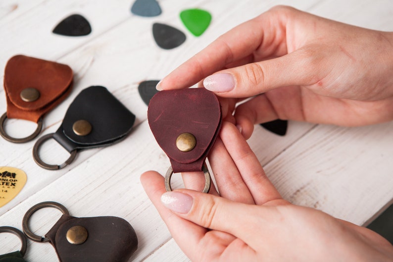 May include: A set of four leather guitar pick holders with keychains. The holders are in brown, black, and burgundy. The keychains are silver.