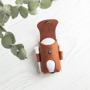 May include: A brown leather golf ball holder with a snap closure, holding two golf balls and two tees. The holder is on a white wooden surface, with green leaves in the background. The holder is a stylish accessory for golfers.