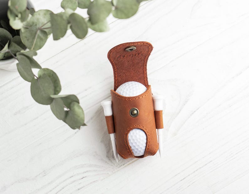 May include: A brown leather golf ball holder with a snap closure, holding two golf balls and two tees. The holder is on a white wooden surface, with green leaves in the background. The holder is a stylish accessory for golfers.