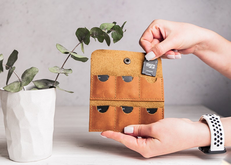 K&ouml;nnte beinhalten: Ein braunes Lederetui mit sechs Steckpl&auml;tzen f&uuml;r Speicherkarten. Das Etui ist ge&ouml;ffnet und eine Person h&auml;lt einen Micro-SD-Kartenadapter in der Hand.