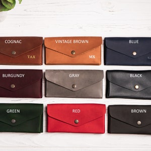 May include: Nine leather wallets in different colors, including cognac, vintage brown, blue, burgundy, gray, black, green, red, and brown. Each wallet has a gold button and is labeled with the color name.