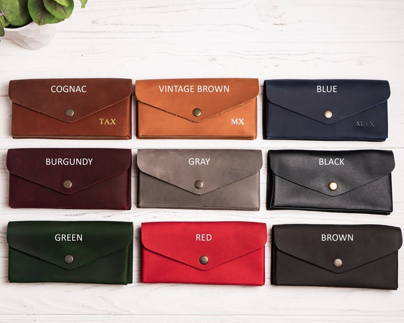 May include: Nine leather wallets in different colors, including cognac, vintage brown, blue, burgundy, gray, black, green, red, and brown. Each wallet has a gold button and is labeled with the color name.
