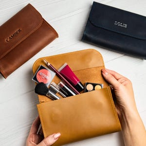 Puede incluir: Un neceser de maquillaje de cuero marrón claro está abierto, revelando varios cosméticos y brochas. También hay dos estuches de cuero adicionales en marrón y azul marino. El estuche marrón tiene el nombre "CAMBER" grabado, y el azul marino "LOLIE".