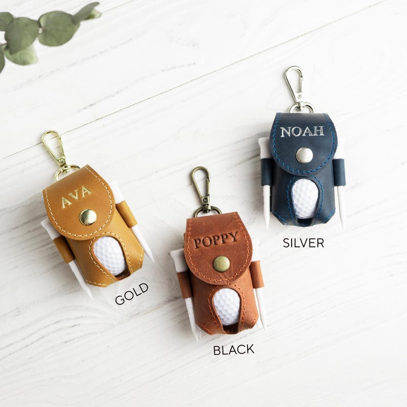 May include: Three leather golf ball holders in gold, brown, and navy blue, each with a golf ball and tees. The gold holder is labeled "AVA", the brown "POPPY", and the navy blue "NOAH". The holders have a snap closure and a metal clip.