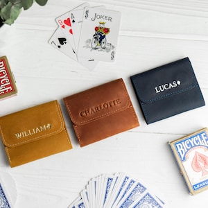 May include: Three leather wallets in mustard, brown, and navy blue, each with a name embossed on the front. Playing cards and two Bicycle card decks are scattered around the wallets on a white wooden surface. The wallets are closed.