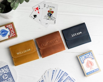 Può includere: Tre portafogli in pelle color senape, marrone e blu navy, ciascuno con un nome impresso sul davanti. Carte da gioco e due mazzi di carte Bicycle sono sparsi intorno ai portafogli su una superficie di legno bianco. I portafogli sono chiusi.