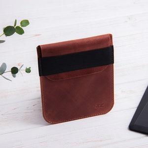 Puede incluir: Una funda de cuero marr&oacute;n con una banda el&aacute;stica negra para un lector electr&oacute;nico. La funda tiene un bolsillo en la parte delantera y las iniciales "XK" est&aacute;n grabadas en la parte inferior.