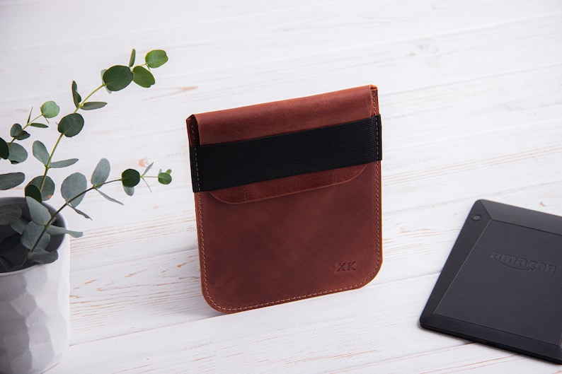 Puede incluir: Una funda de cuero marr&oacute;n con una banda el&aacute;stica negra para un lector electr&oacute;nico. La funda tiene un bolsillo en la parte delantera y las iniciales "XK" est&aacute;n grabadas en la parte inferior.