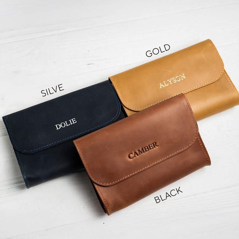 May include: Four leather wallets in navy blue, gold, and brown. Each wallet has a flap closure and is embossed with a name. The wallets are labeled "SILVE", "GOLD", and "BLACK". The navy wallet says "DOLIE", the gold wallet says "ALYSON", and the brown wallet says "CAMBER".