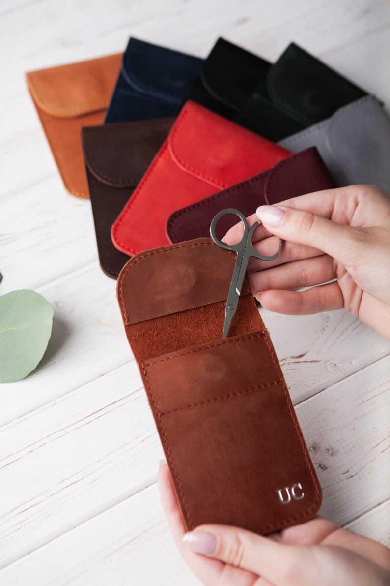 May include: A brown leather case with a flap that opens to reveal a small pair of silver scissors. The case has a single initial, "UC", embossed on the front.