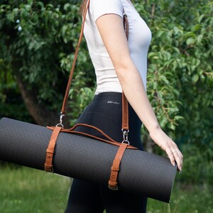 May include: A rolled-up black yoga mat with brown leather straps and a shoulder strap. The mat is secured with two leather straps and buckles. The shoulder strap is also brown leather. The background is a blurred green outdoor scene.