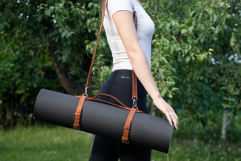 May include: A rolled-up black yoga mat with brown leather straps and a shoulder strap. The mat is secured with two leather straps and buckles. The shoulder strap is also brown leather. The background is a blurred green outdoor scene.