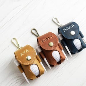May include: Three personalized leather golf ball holders in tan, brown, and navy blue. Each holder has a golf ball and tees. The holders are labeled with the names "AVA", "POPPY", and "NOAH". Each holder has a silver-colored clip.