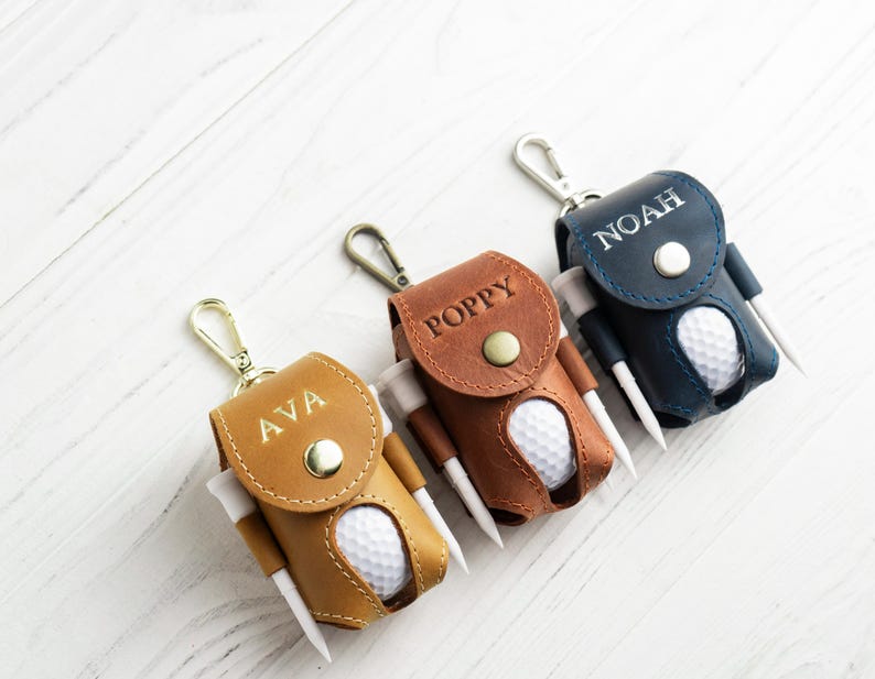 May include: Three personalized leather golf ball holders in tan, brown, and navy blue. Each holder has a golf ball and tees. The holders are labeled with the names "AVA", "POPPY", and "NOAH". Each holder has a silver-colored clip.
