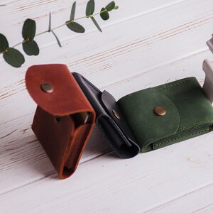 Pu&ograve; includere: Tre portafogli in pelle marrone, nera e verde, ciascuno con chiusura a scatto. I portafogli sono disposti su una superficie di legno bianco, con un rametto di eucalipto e una statuina decorativa a forma di cervo sullo sfondo.