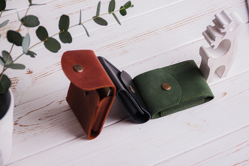 Pu&ograve; includere: Tre portafogli in pelle marrone, nera e verde, ciascuno con chiusura a scatto. I portafogli sono disposti su una superficie di legno bianco, con un rametto di eucalipto e una statuina decorativa a forma di cervo sullo sfondo.