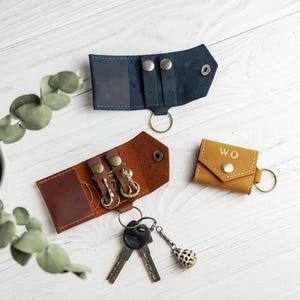 May include: Three leather key organizers in navy blue, brown, and tan. The tan organizer is closed and features the letters W.O. The brown and navy blue organizers are open, showing internal metal snaps and key rings. One set of keys is attached.