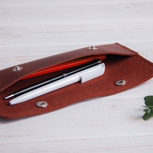 May include: A brown leather pencil case with a flap opening, lined with red felt. Two pens and a red pencil are inside the case.