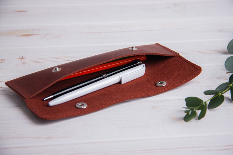 May include: A brown leather pencil case with a flap opening, lined with red felt. Two pens and a red pencil are inside the case.