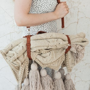 May include: A beige knitted blanket with a brown leather strap and shoulder strap. The blanket has a decorative pattern and is adorned with multiple beige tassels. The blanket is rolled up and carried over the shoulder.