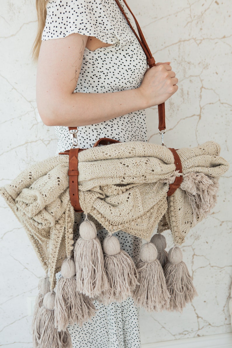 May include: A beige knitted blanket with a brown leather strap and shoulder strap. The blanket has a decorative pattern and is adorned with multiple beige tassels. The blanket is rolled up and carried over the shoulder.