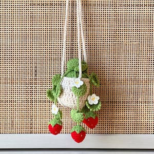 May include: A crocheted strawberry-shaped hanging planter with a cream-colored pouch and green leaves. The planter has three red strawberries hanging from it.