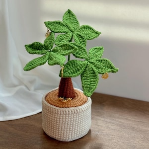 May include: A crocheted plant in a white pot. The plant has green leaves and a brown trunk. The pot is beige and has a textured design. The text "LittleMoreRich" is at the bottom of the image.